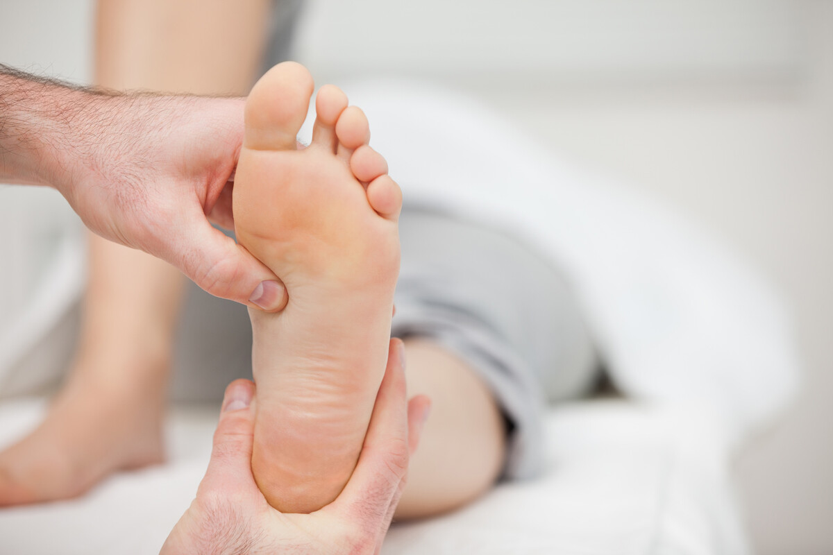 Mani di un operatore sorreggono la pianta del piede nudo di una persona sdraiata su un lettino con lenzuolo bianco