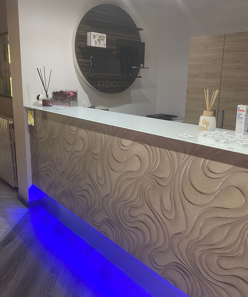 Bancone reception con fronte beige a onde, piano bianco e LED blu, dietro pannello tondo in legno con scritta FISIO